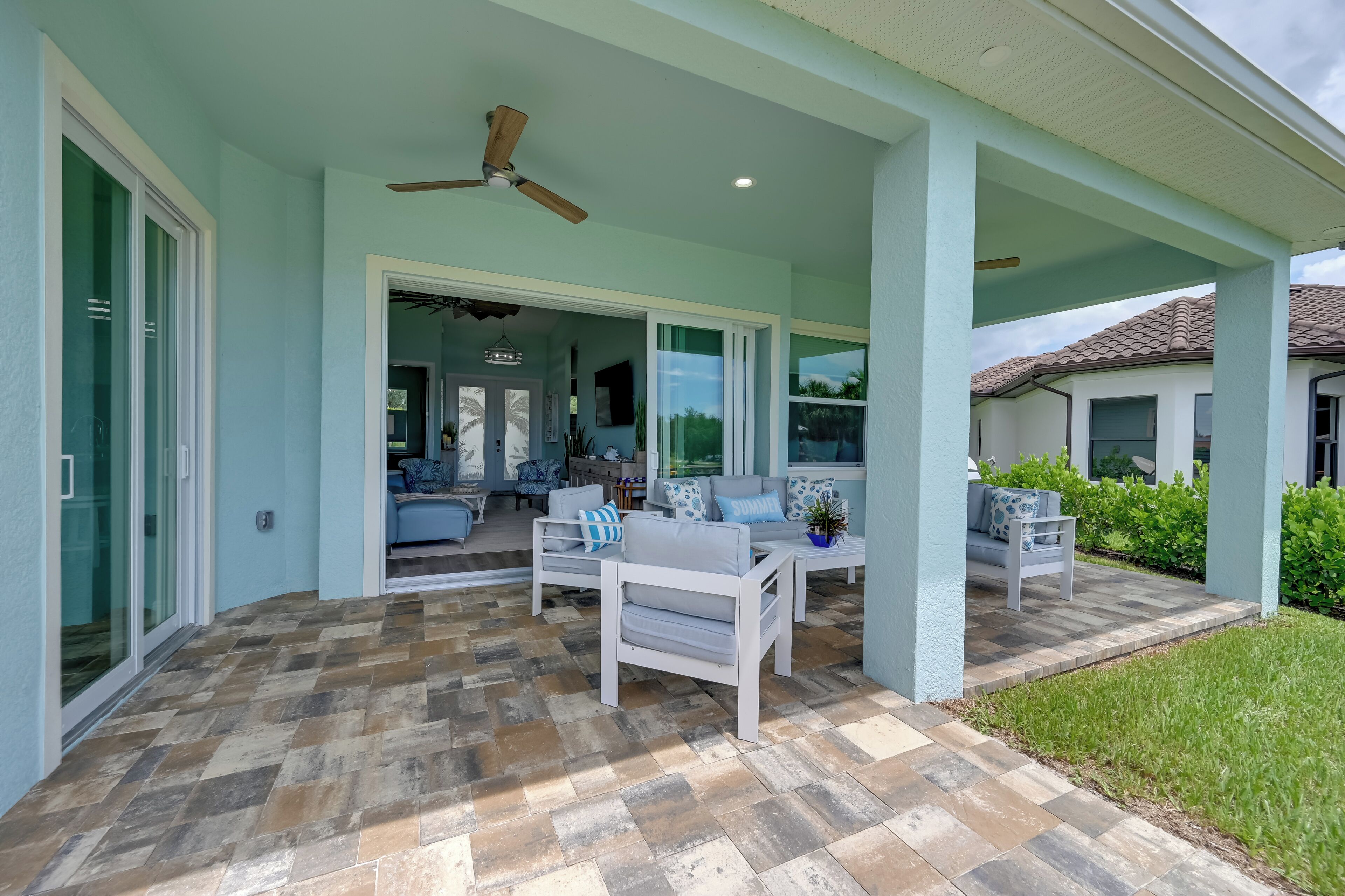 Un patio couvert avec un ventilateur de plafond, des sièges d’extérieur et une vue sur l’intérieur de la maison.