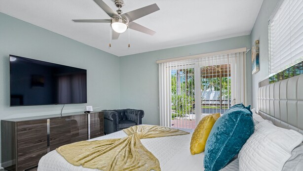 A bedroom with a bed, a TV, a ceiling fan, a desk, and a window with a view of greenery.