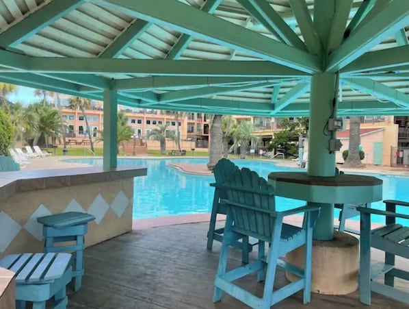 Un espace piscine couvert, équipé d’une table de billard et de chaises, donnant sur une piscine et un ensemble de bâtiments.