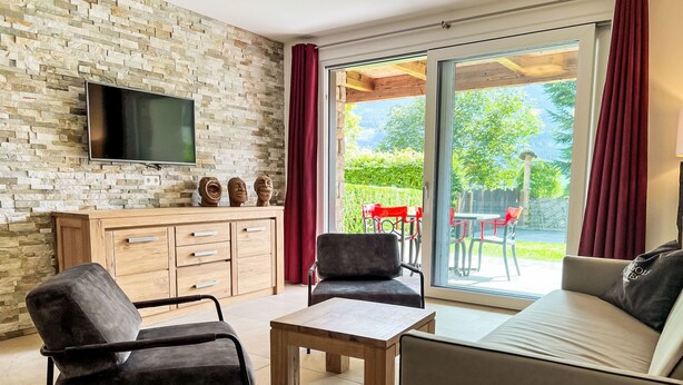 Un salon moderne avec un mur en pierre, un téléviseur à écran plat, un meuble en bois et une porte-fenêtre coulissante donnant sur un espace extérieur agrémenté de chaises rouges.