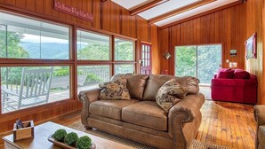 A cozy living room with a brown leather sofa, a red armchair, and a wooden coffee table. Large windows offer a view of a balcony and mountains. - Mountain Dreams- Peaceful, Charming, Pet Friendly (Maggie Valley)