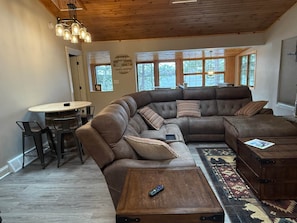 A cozy living room with a large L-shaped sectional sofa, a round coffee table, and a dining area with a table and chairs. - Lost in the Woods Lodge (Saint Germain)