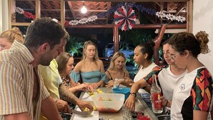 Un grupo de personas reunidas alrededor de una mesa con comida y bebidas, en una habitación con techo de madera y una ventana decorada.