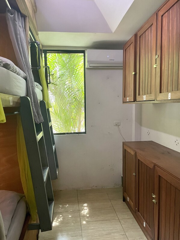 Habitación pequeña con literas, ventana que da a follaje verde, un armario de madera y un equipo de aire acondicionado.