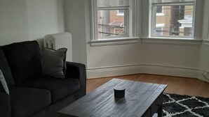 A living room with a dark sofa, a wooden coffee table, and a black and white rug. There are two windows with blinds, and a radiator is visible on the wall.