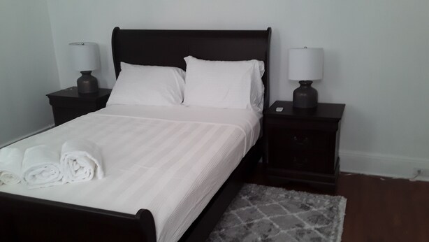 A neatly made bed with white linens and rolled towels, two bedside tables with lamps, and a patterned rug on a wooden floor.