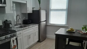 A kitchen with white cabinets, a stainless steel refrigerator, a stove, and a sink. There is a window with blinds and a table with chairs.
