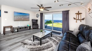 Ein modernes Wohnzimmer mit großem Fenster, Deckenventilator, schwarzem Ledersofa, Couchtisch und Blick auf den Strand.