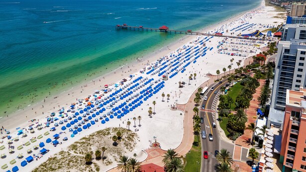 Eine Luftaufnahme eines Strandes mit Reihen blauer Sonnenschirme, einem Steg und einer Straße neben einem Hotel.