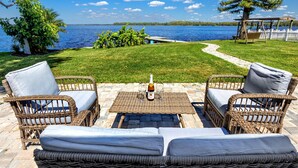 Eine Terrasse mit Korbmöbeln, einem Tisch mit Flasche und Gläsern sowie Blick auf einen See.