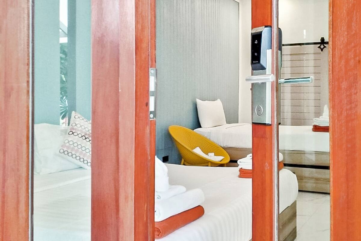 Une chambre d’hôtel moderne avec un lit, une chaise jaune et une salle de bain visible à travers une paroi en verre.