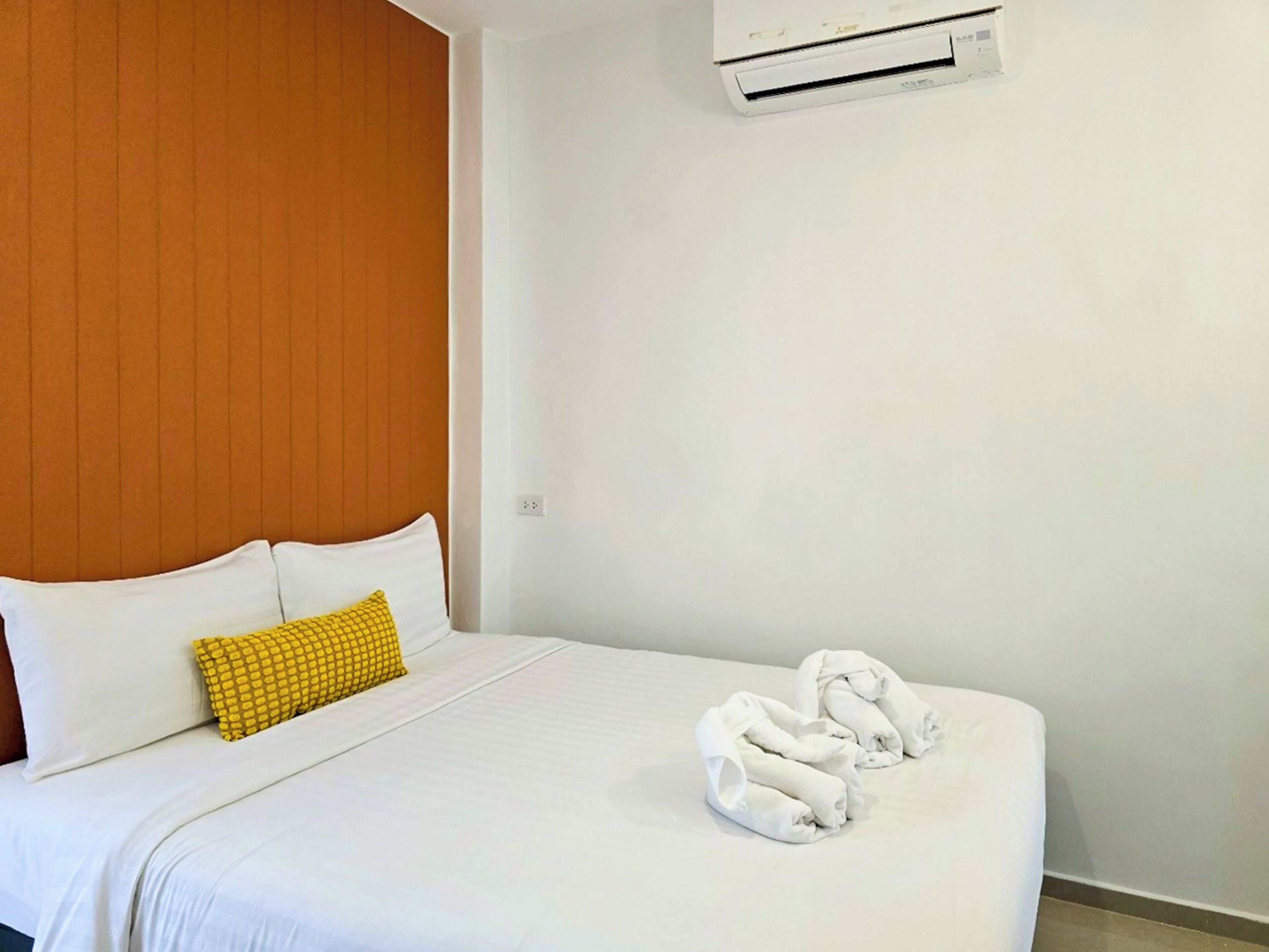 Habitación de hotel con cama, cabecera de madera, pared blanca y equipo de aire acondicionado, además de un almohadón amarillo.