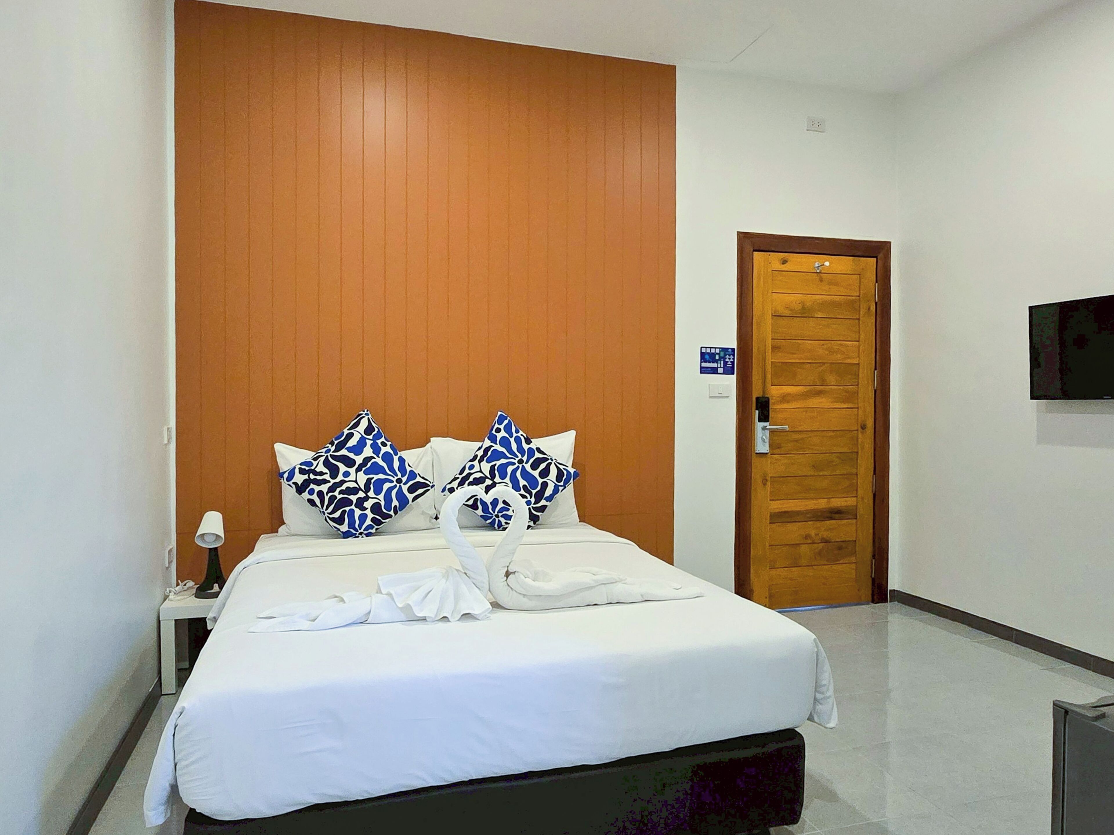 A hotel room with a bed, a wooden door, a TV mounted on the wall, and a nightstand with a lamp.