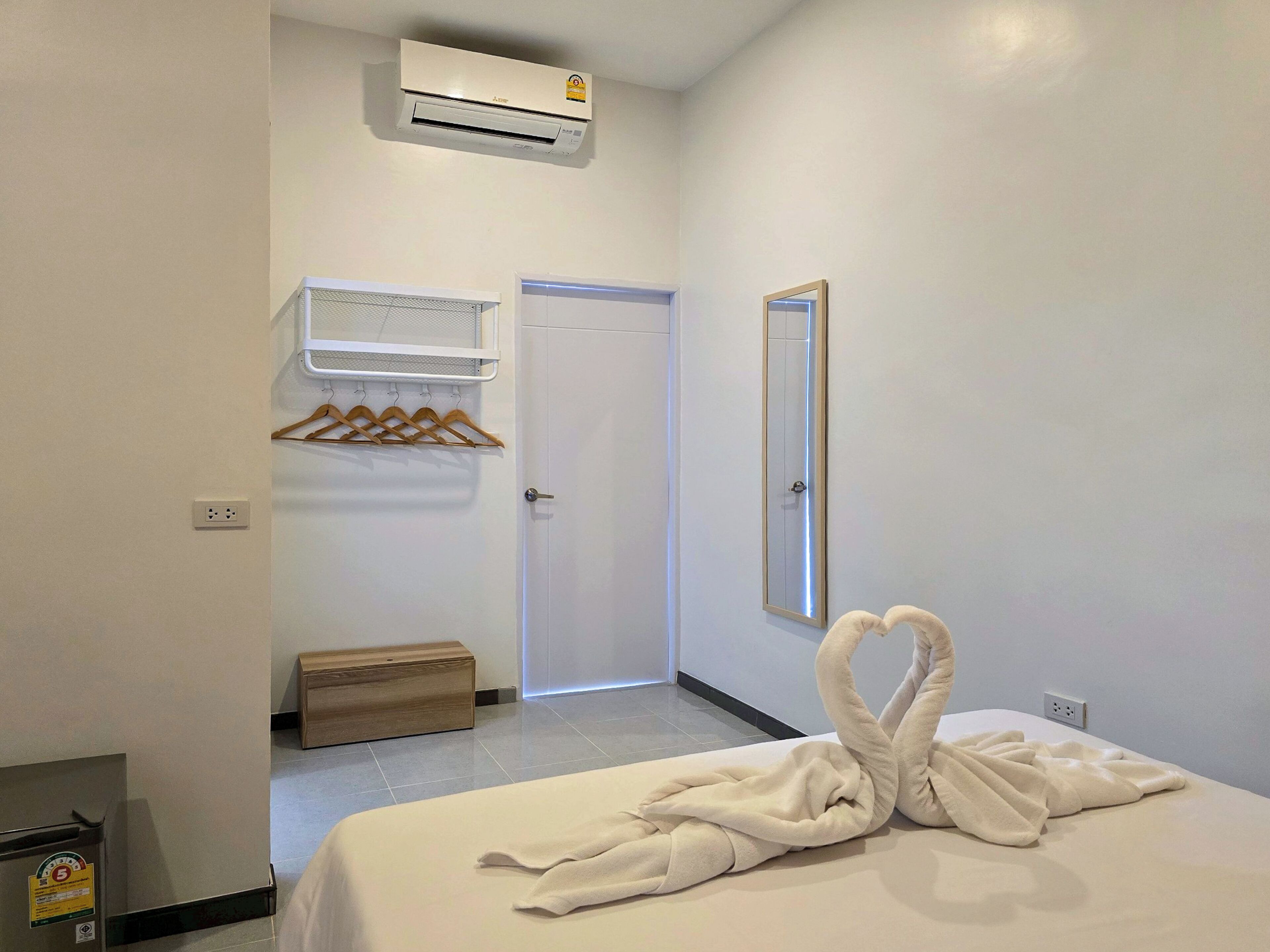 A hotel room with a bed, an air conditioning unit, a mirror, a coat rack, and a wooden bench.
