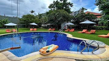 Piscina com espreguiçadeiras e guarda-sóis, cercada por vegetação e edifícios.