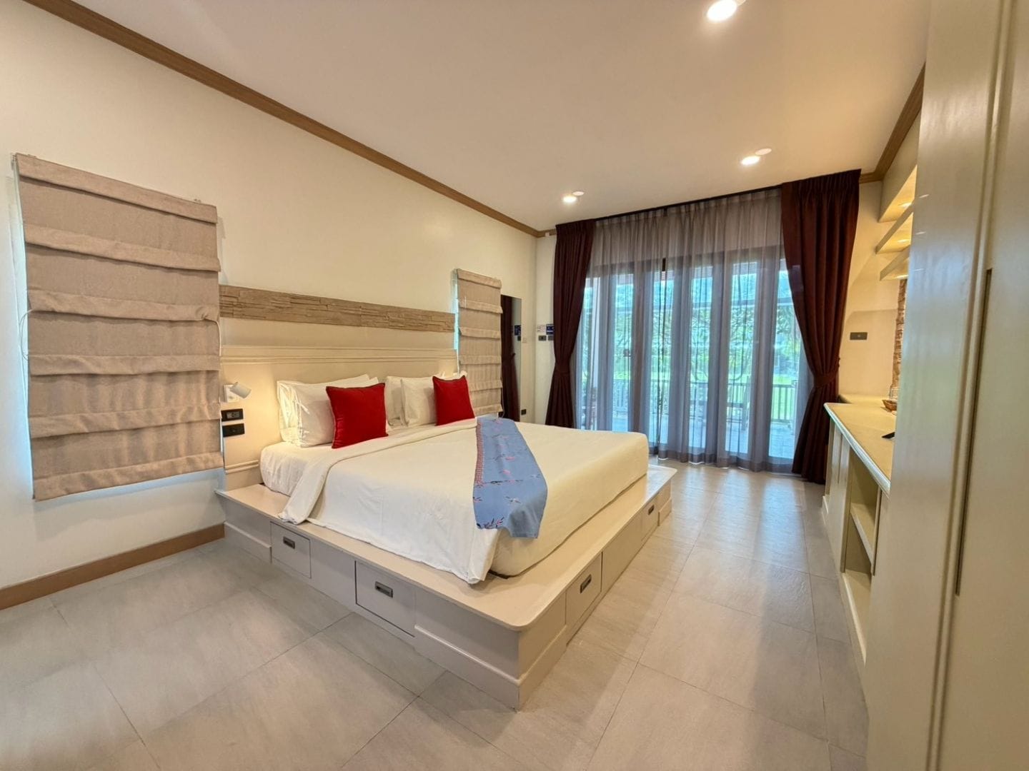 Un dormitorio moderno con una cama grande, cajones de almacenamiento empotrados, una mesita de noche y vistas al exterior a través de amplias ventanas.