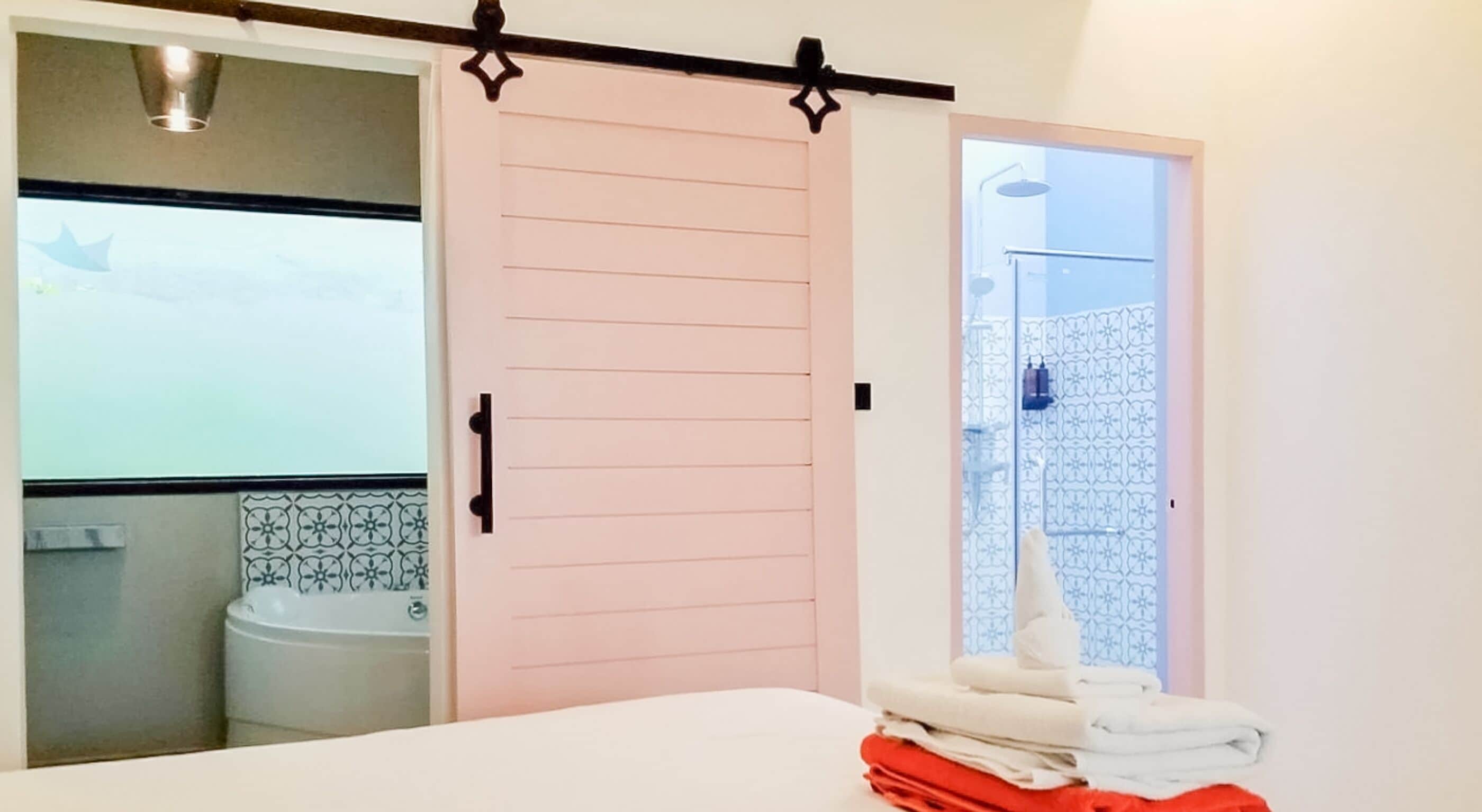 A bedroom with a bed, a pink sliding door, a bathroom with a toilet and sink, and a towel rack with towels.