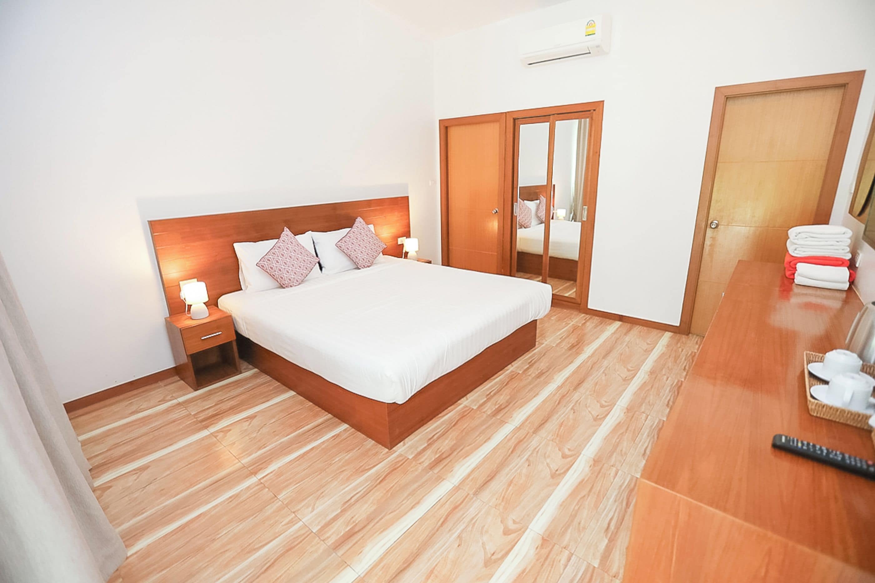 A hotel room with a wooden floor, a bed with white bedding, a nightstand with a lamp, a wardrobe, and a bathroom with towels.
