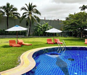 A swimming pool with a blue tiled floor, surrounded by grass and palm trees. There are red lounge chairs and umbrellas.