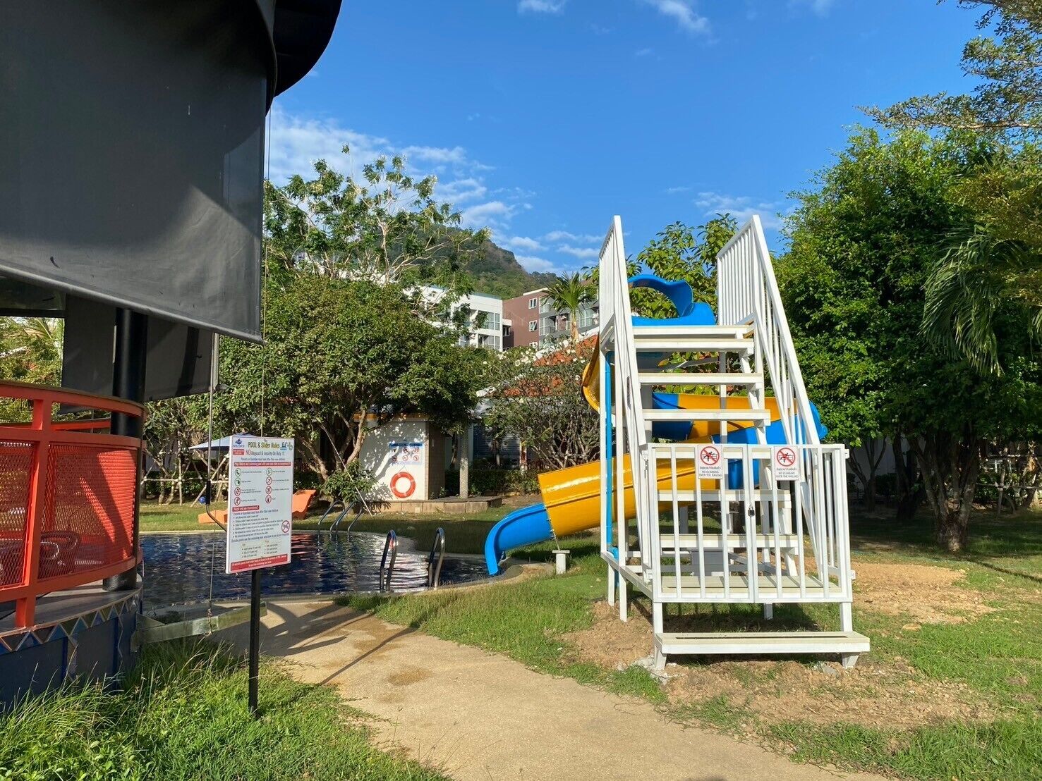 A playground with a slide and climbing structure, surrounded by greenery and a clear sky.