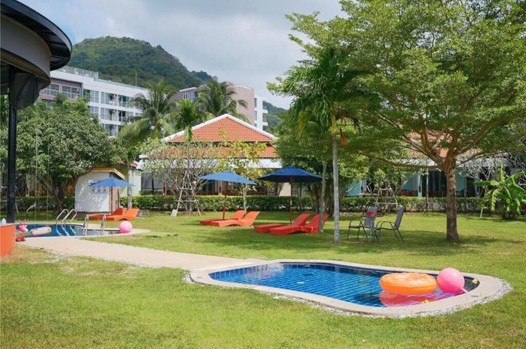 A pool area with lounge chairs, umbrellas, and a small building, surrounded by greenery and buildings in the background.