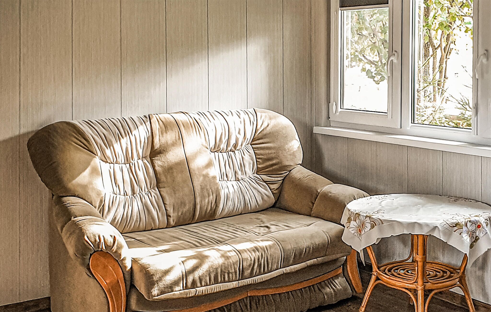 A room with a beige upholstered sofa, a small round table with a white tablecloth, and a window with a view of trees.