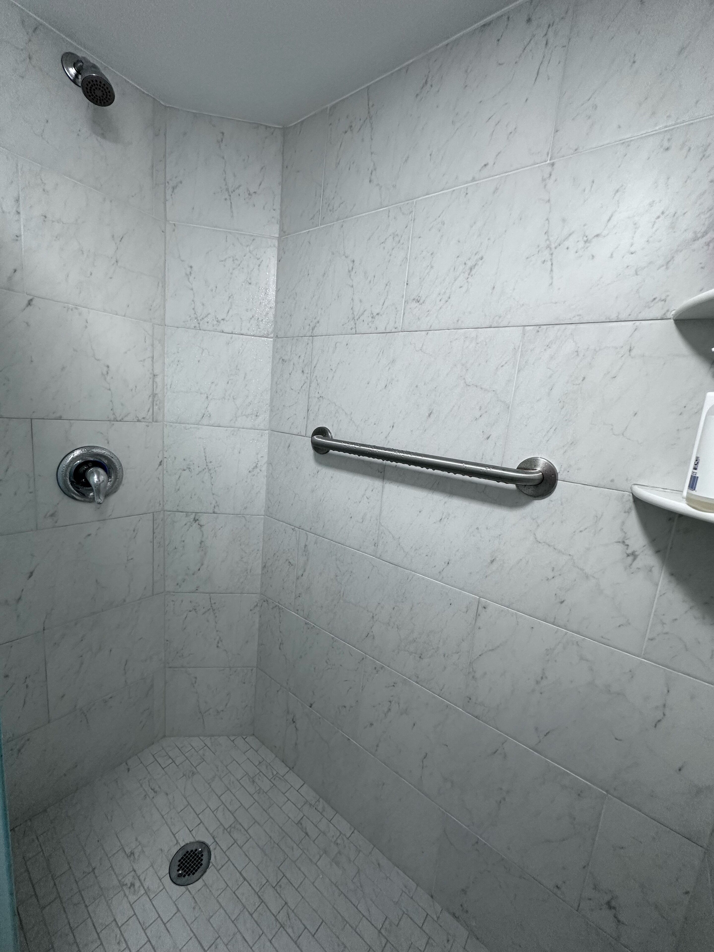 A shower with white marble tiles, a handheld showerhead, a handheld shower bar, and a floor drain.