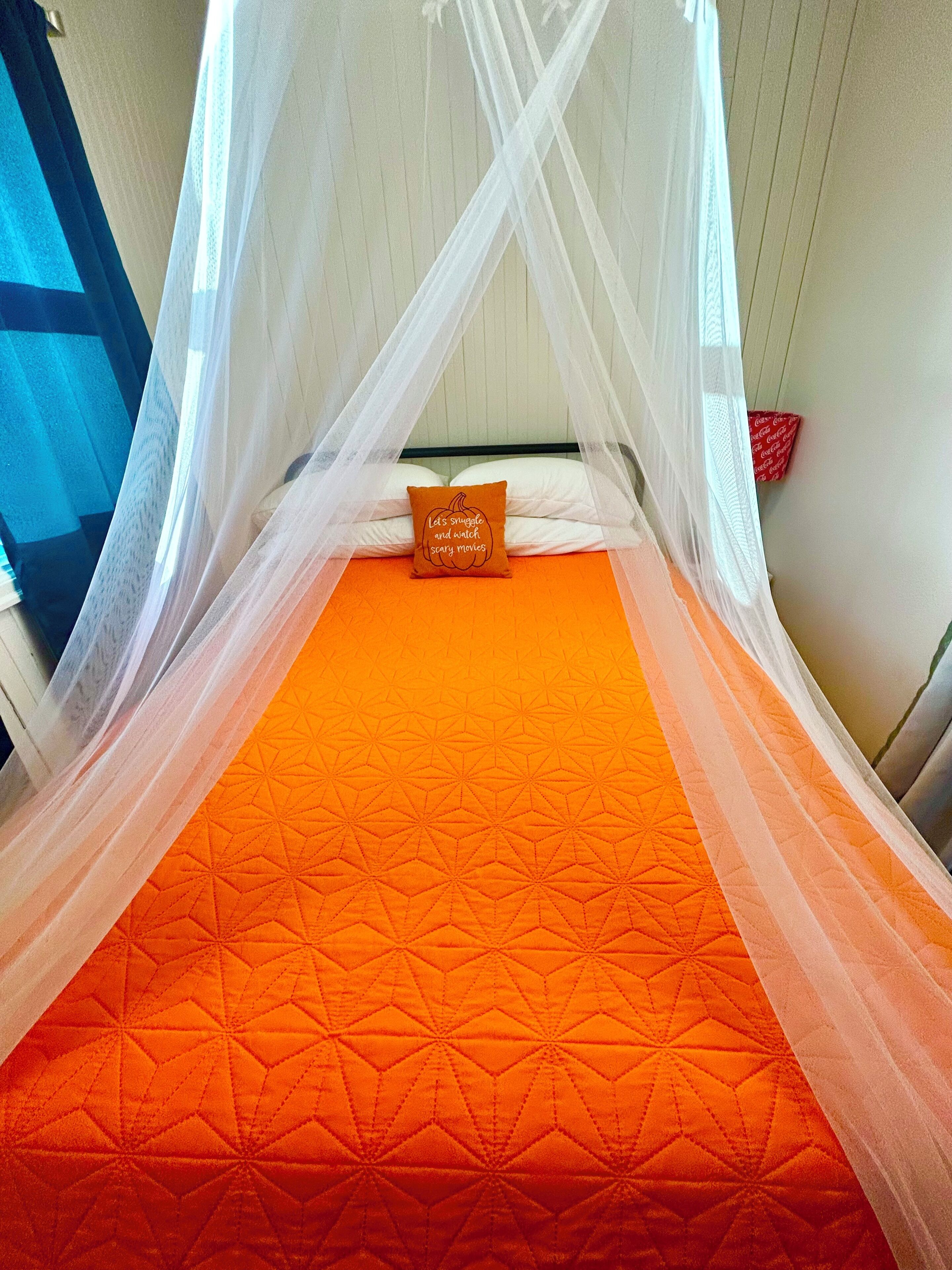 Un lit avec une couette orange à motifs géométriques, des oreillers blancs et un baldaquin.