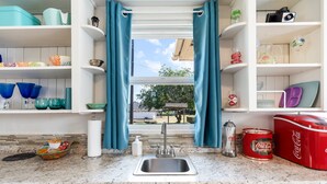 Cozinha com pia, prateleiras com diversos objetos e janela com cortinas.