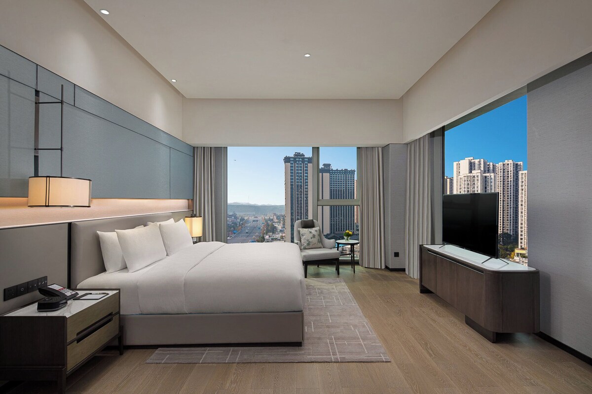 A modern hotel room with a large bed, a TV, a desk, and a view of the cityscape.