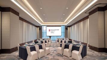 A conference room with a large screen displaying a presentation, arranged tables with chairs, and a carpeted floor.