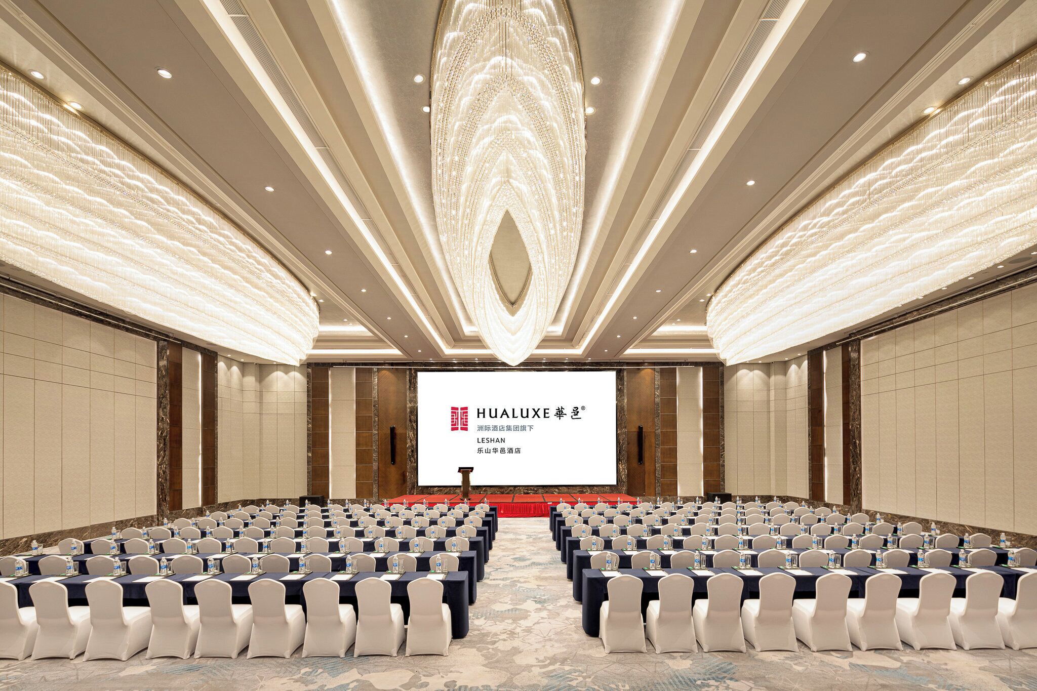 Une salle de conférence avec des rangées de tables et de chaises, un grand écran à l’avant et un plafond décoratif.