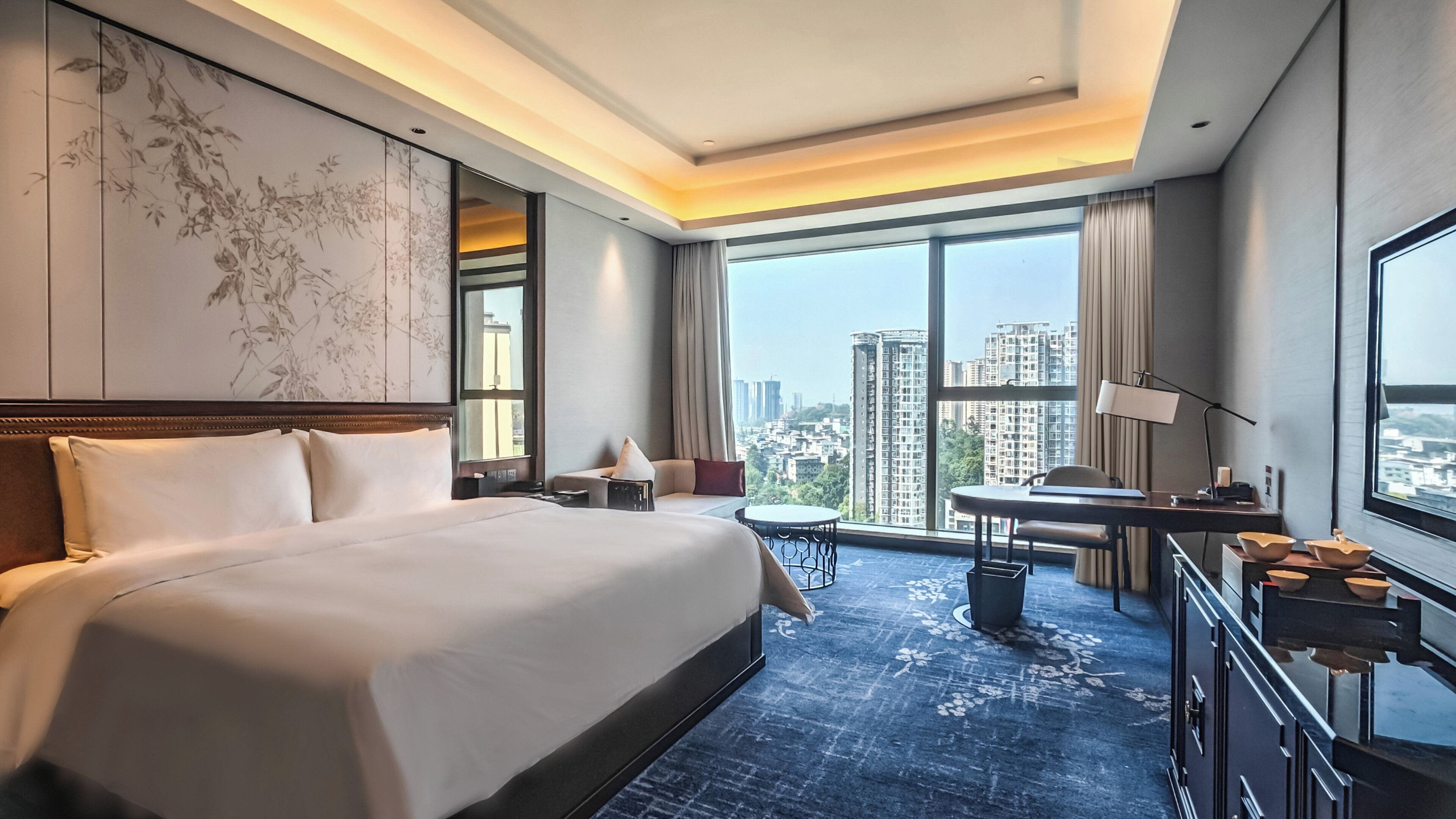 Habitación de hotel con una cama grande, un escritorio, una silla, una mesita y vistas a la ciudad a través de amplios ventanales.