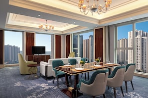 A spacious hotel room with a large dining table, comfortable seating, a flat-screen TV, and a city view.