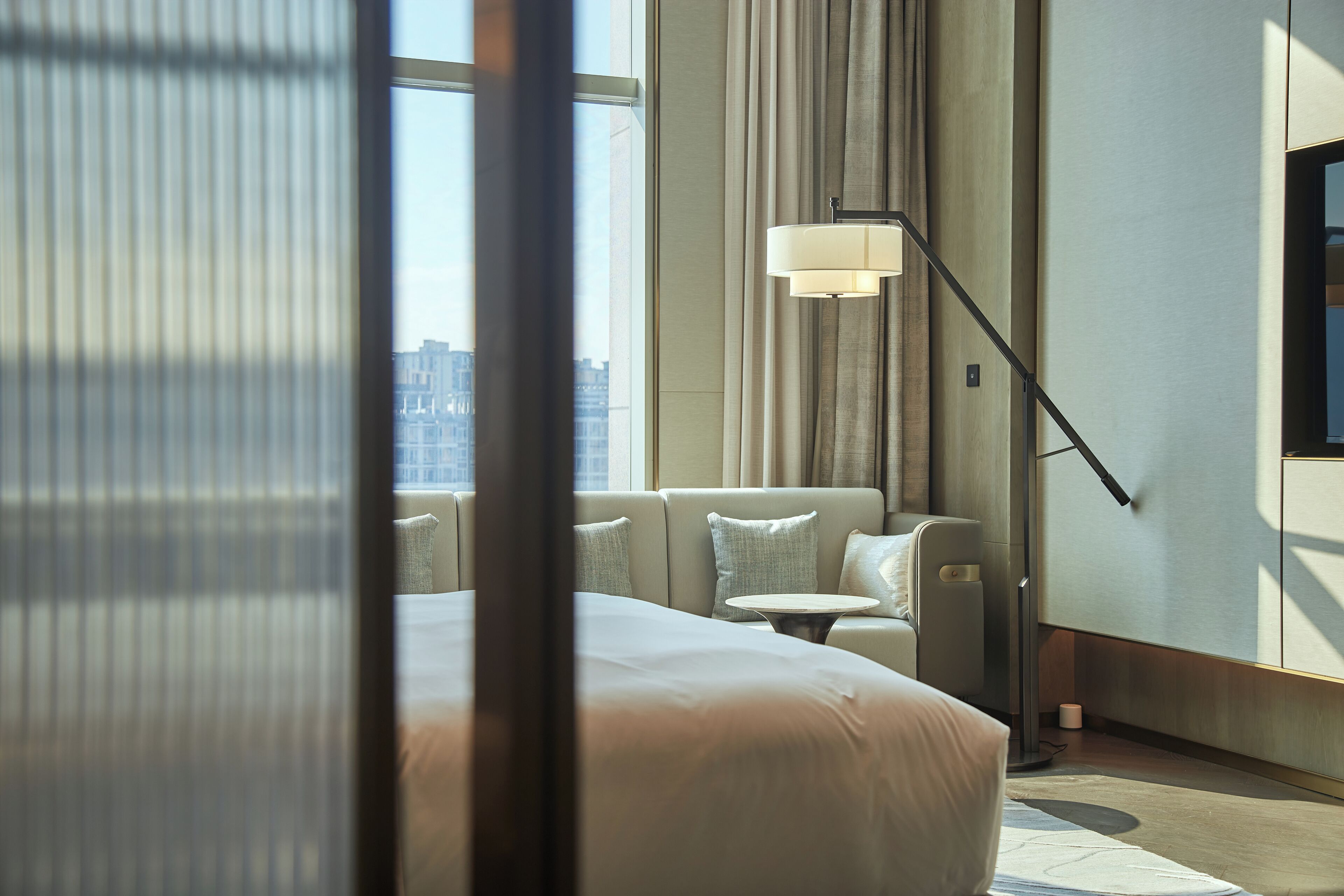 Habitación de hotel moderna con una cama grande, un sofá, una lámpara de pie y vistas a la ciudad a través de amplios ventanales.