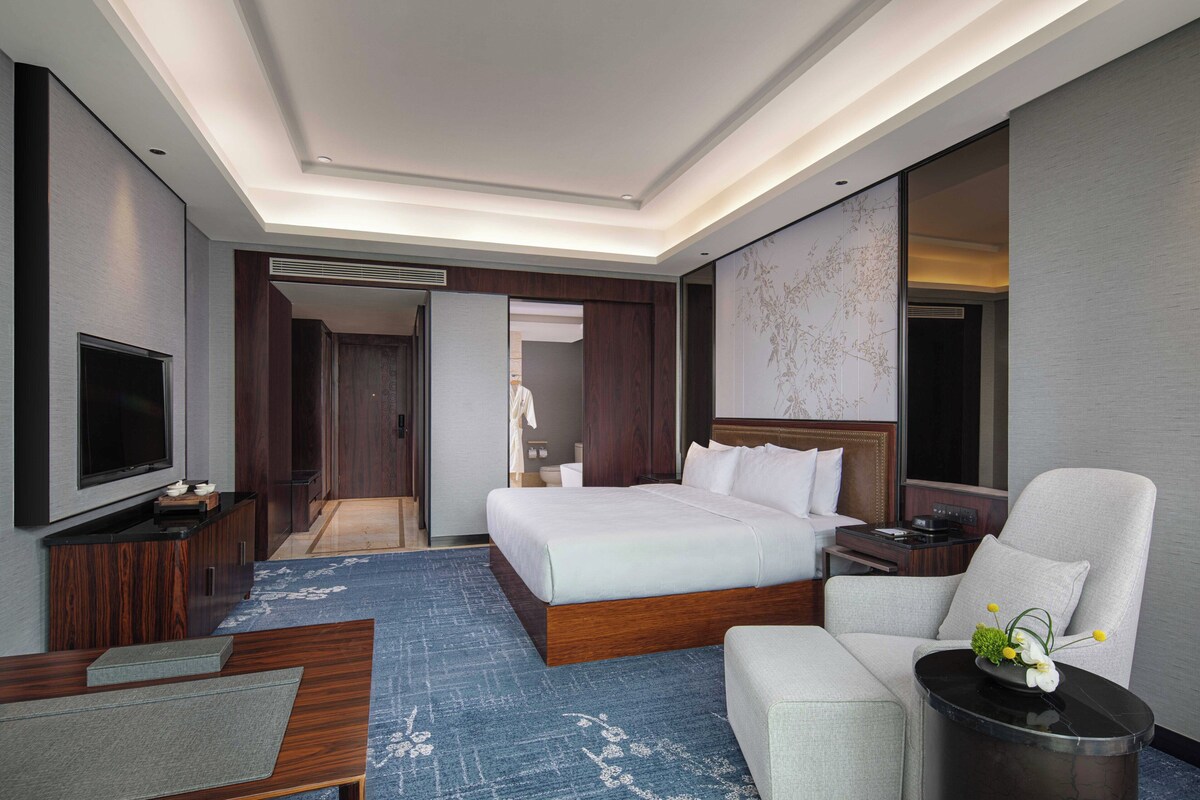 A modern hotel room with a large bed, a flat-screen TV, a sofa, and a small table with a vase of flowers.