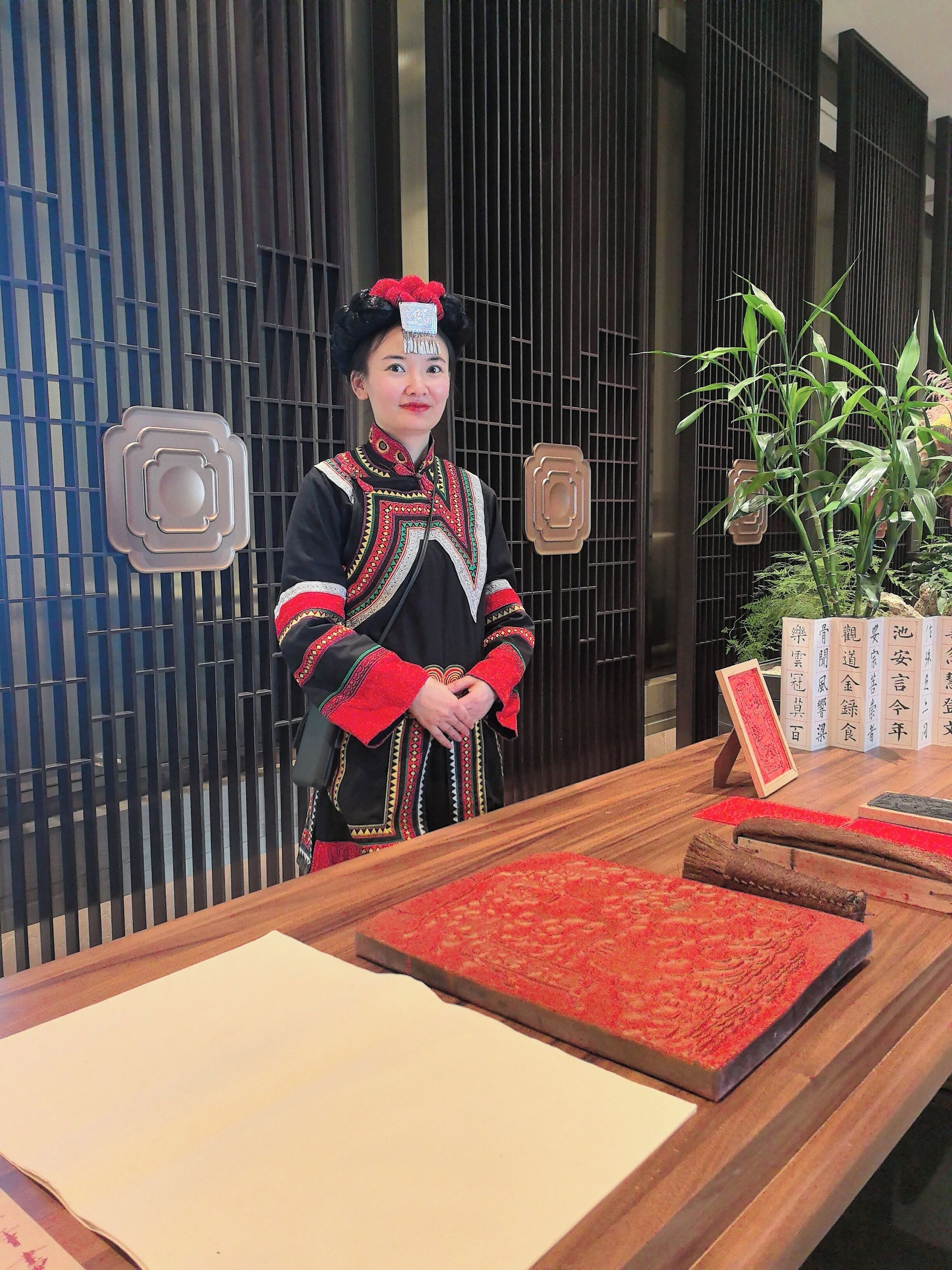 A person in traditional attire standing at a reception desk with a wooden surface, a red patterned mat, and a potted plant in the background.