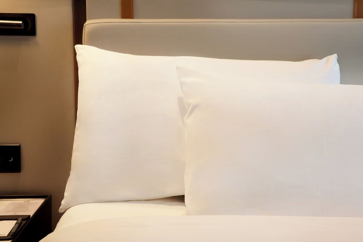 A hotel room with a bed, white pillows, a bedside table with a lamp, and a wall-mounted phone.