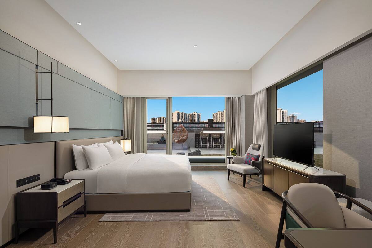 A modern hotel room with a large bed, a desk, a TV, and a view of the city.