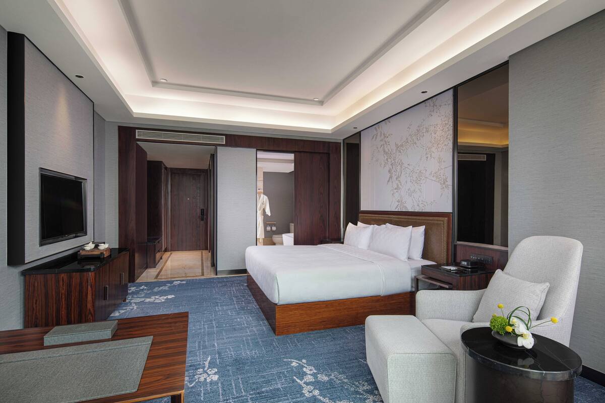 A modern hotel room with a large bed, a flat-screen TV, a sofa, and a small table with a vase of flowers.