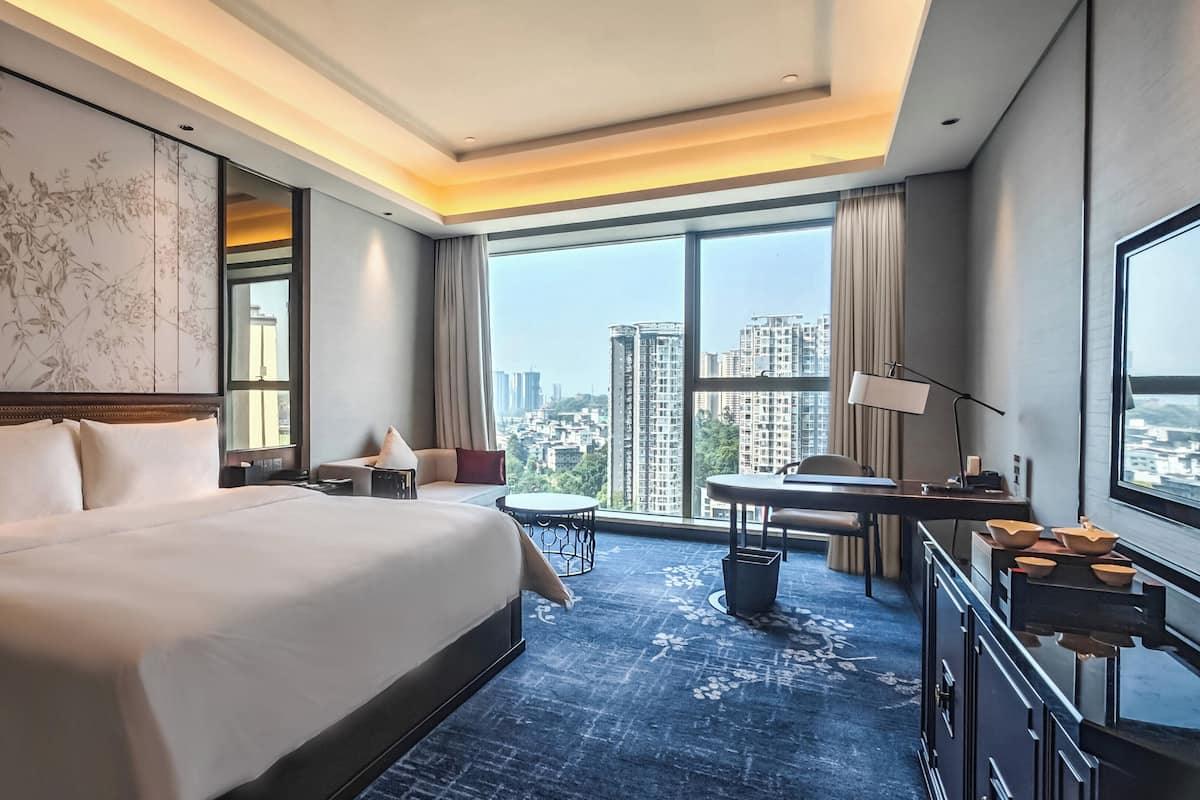 A hotel room with a large bed, a desk, a chair, a small table, and a view of the cityscape through large windows.
