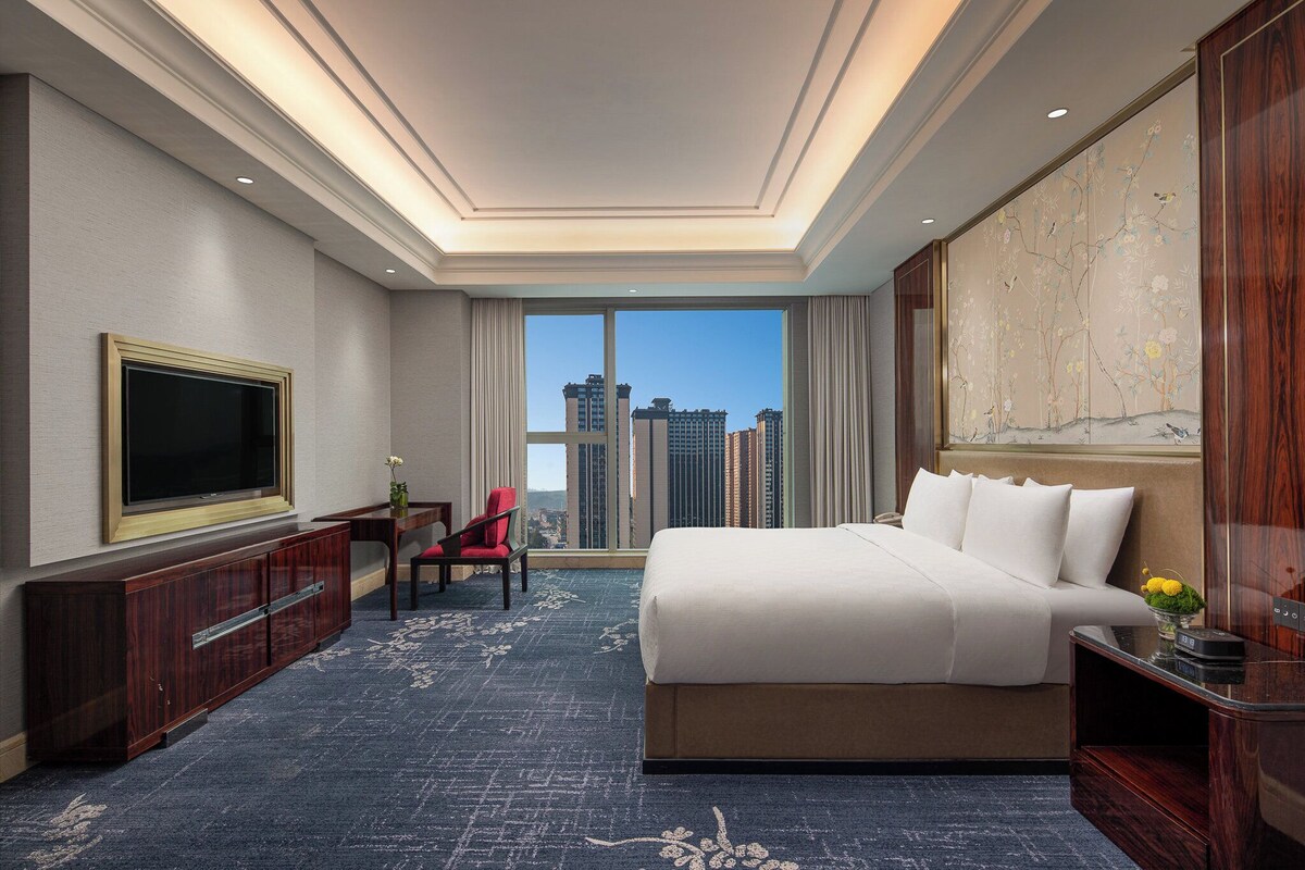 A hotel room with a large bed, a TV, a desk, and a view of the cityscape.
