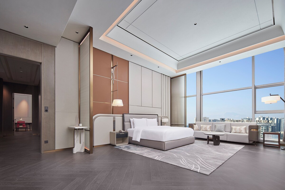 A modern hotel room with a large bed, a sofa, a coffee table, and a view of the city.