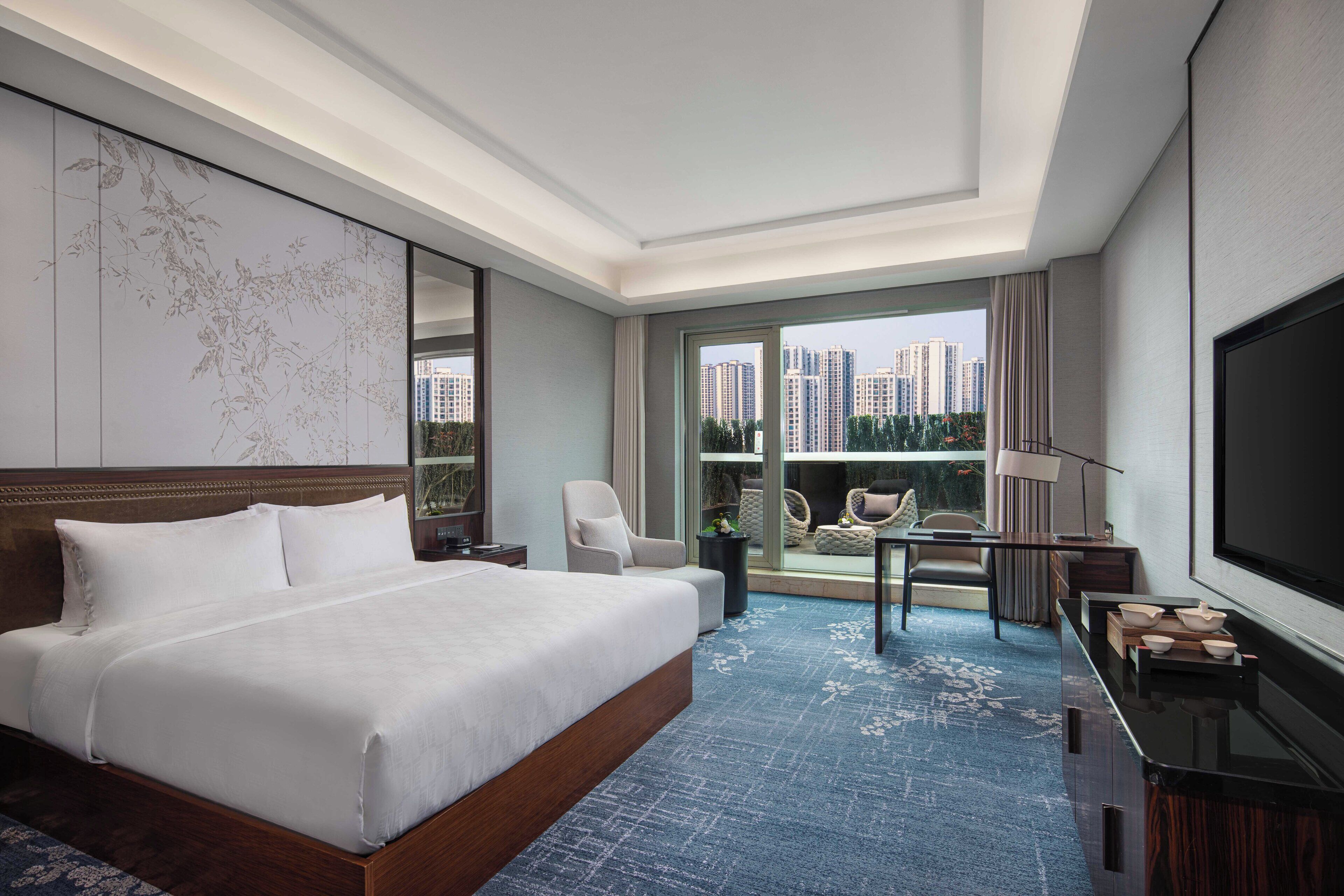 Habitación de hotel con una cama grande, televisión, balcón con vista a edificios altos y zona de estar con mesa y sillas.