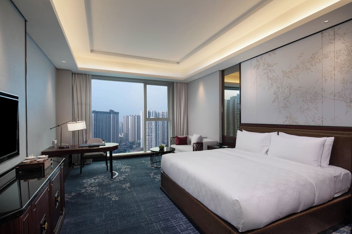 A hotel room with a large bed, a desk with a chair, a TV, and a view of the cityscape through large windows.