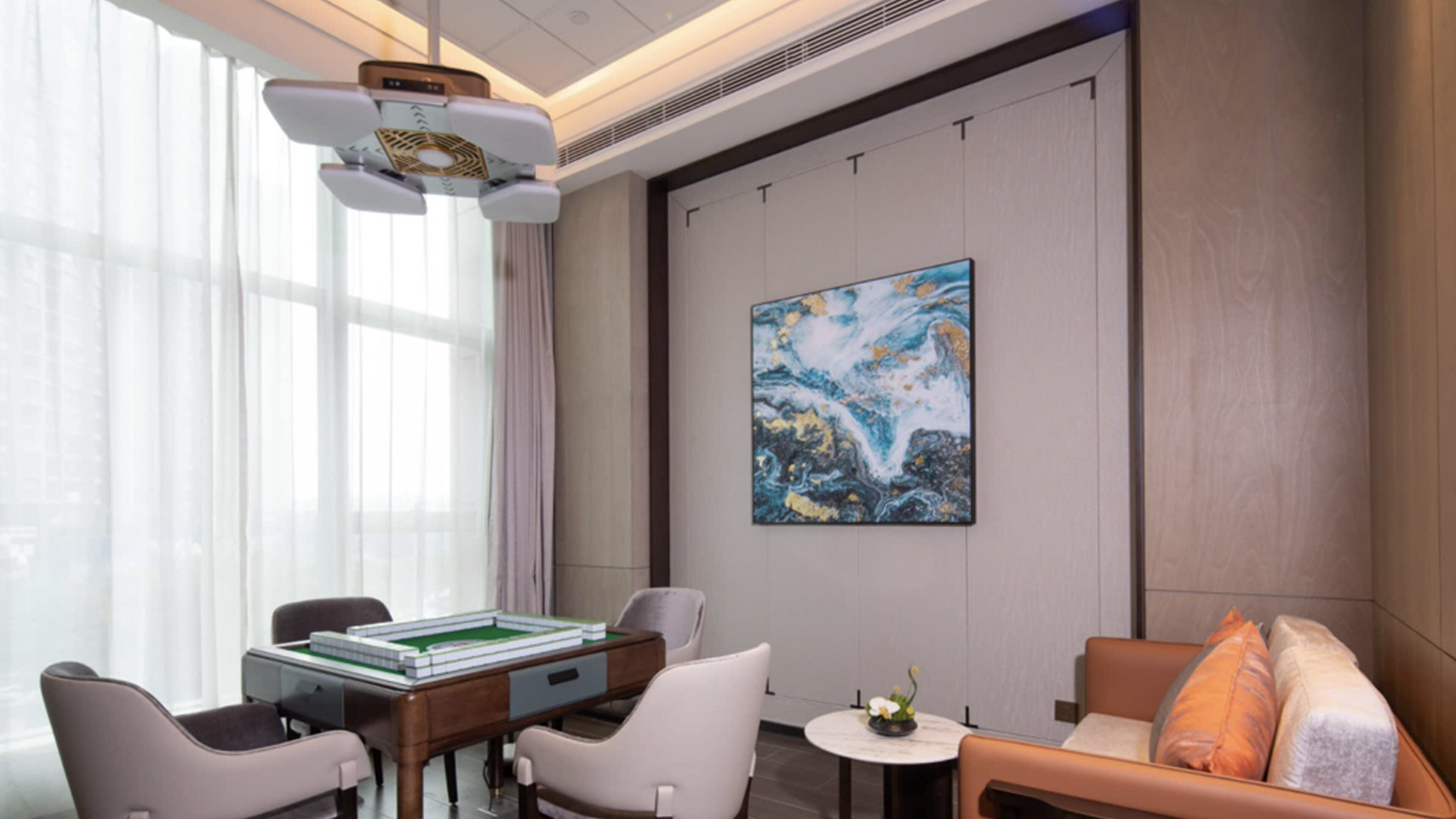 Ein modernes Hotelzimmer mit einem Spieltisch mit grüner Filzauflage, zwei Ledersesseln, einer Couch mit Kissen, einem Bild an der Wand und einem großen Fenster mit durchsichtigen Vorhängen.