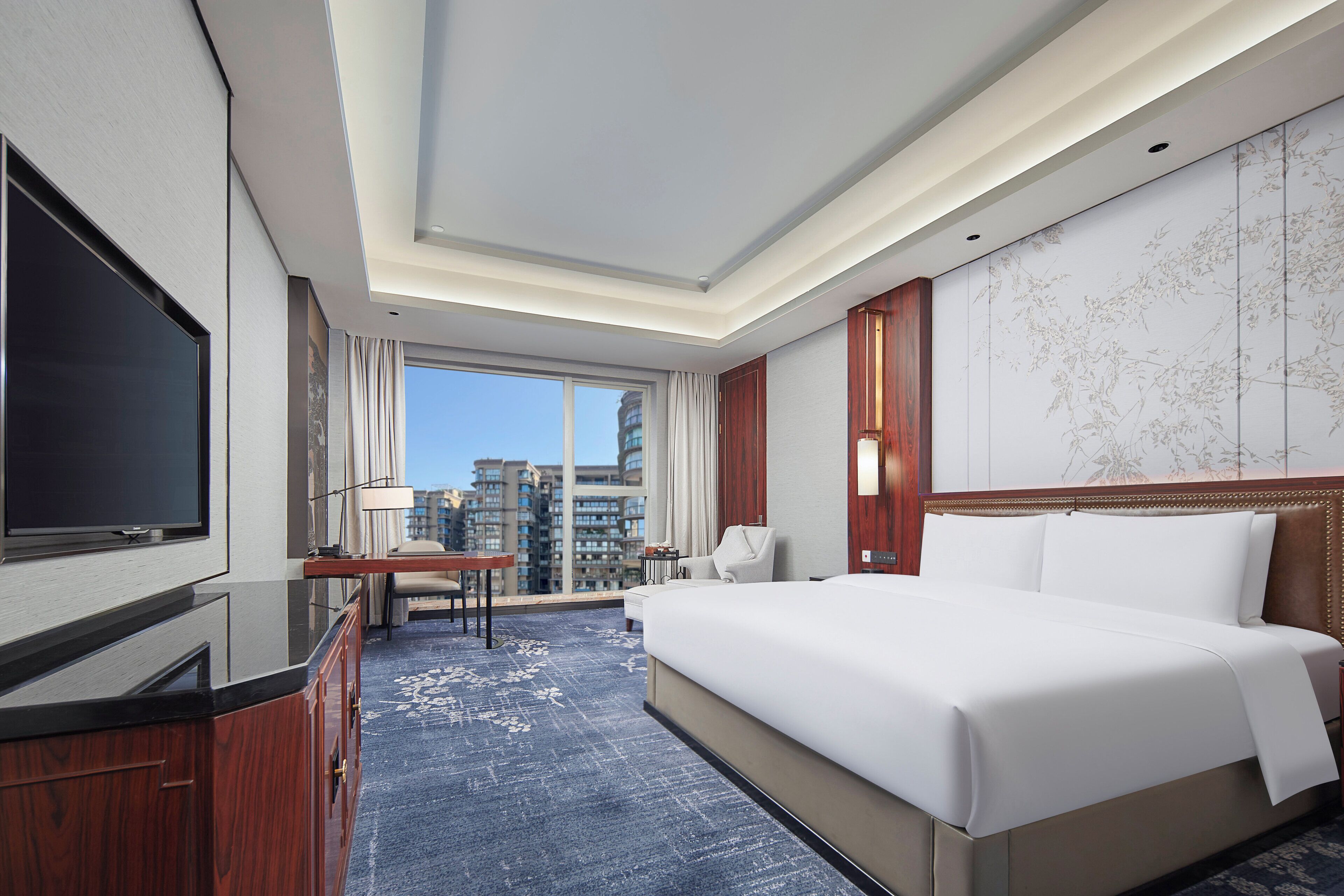 Hotelkamer met een groot bed, een flatscreen televisie, een bureau met een lamp, uitzicht op de stad door een groot raam, en een decoratief wandpaneel.