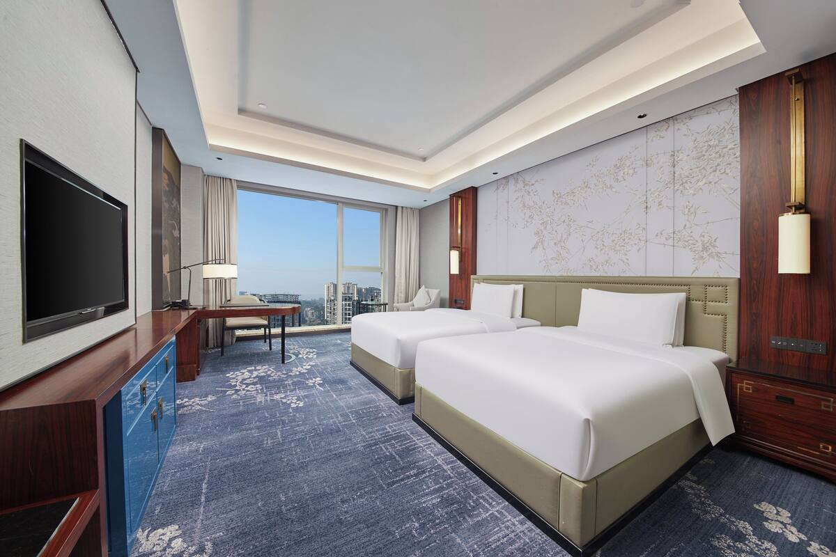 A hotel room with two beds, a desk, a TV, and a large window with a city view.