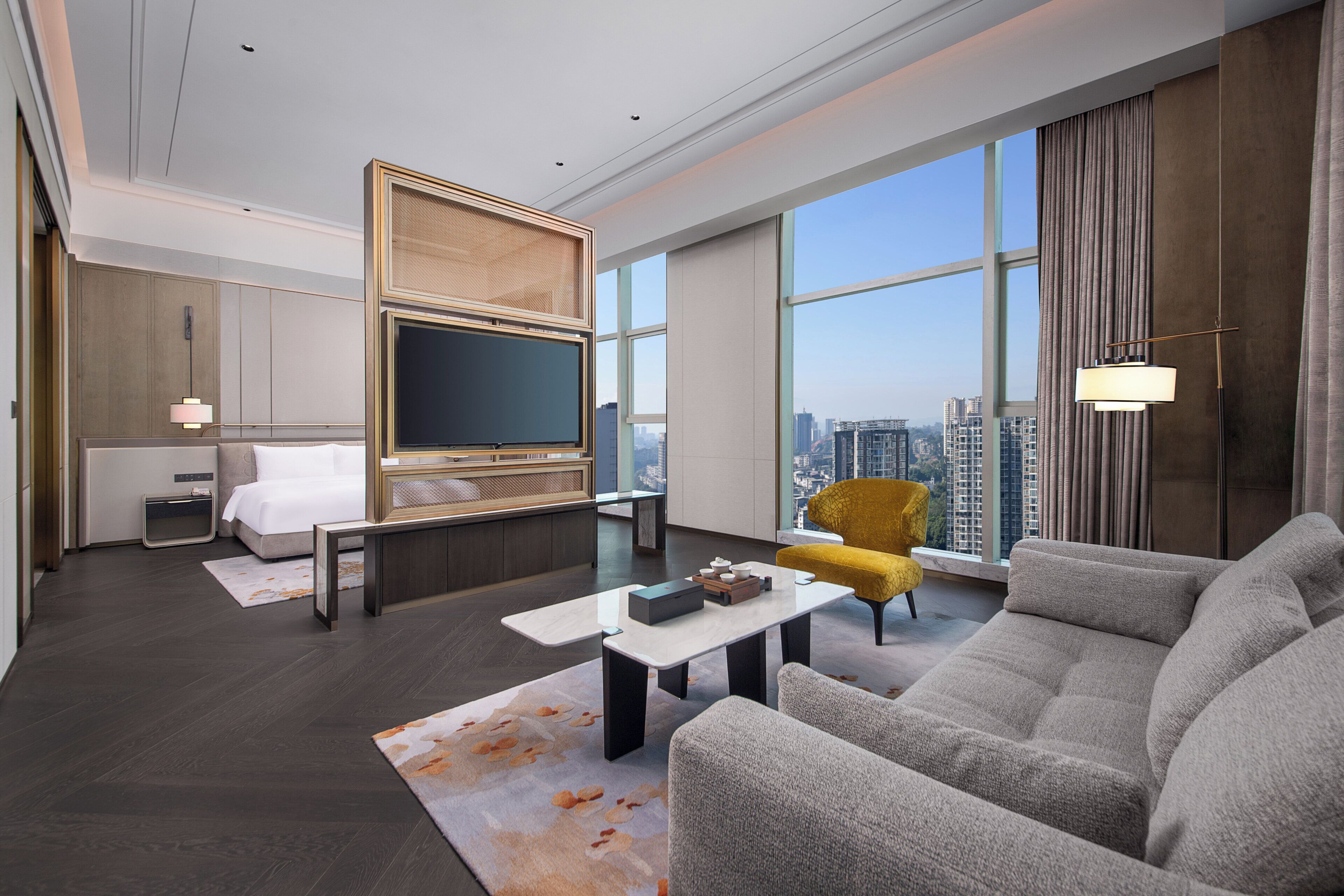 Una habitación de hotel moderna con una cama grande, un televisor de pantalla plana, un sofá, un sillón amarillo y vistas a la ciudad a través de grandes ventanales.