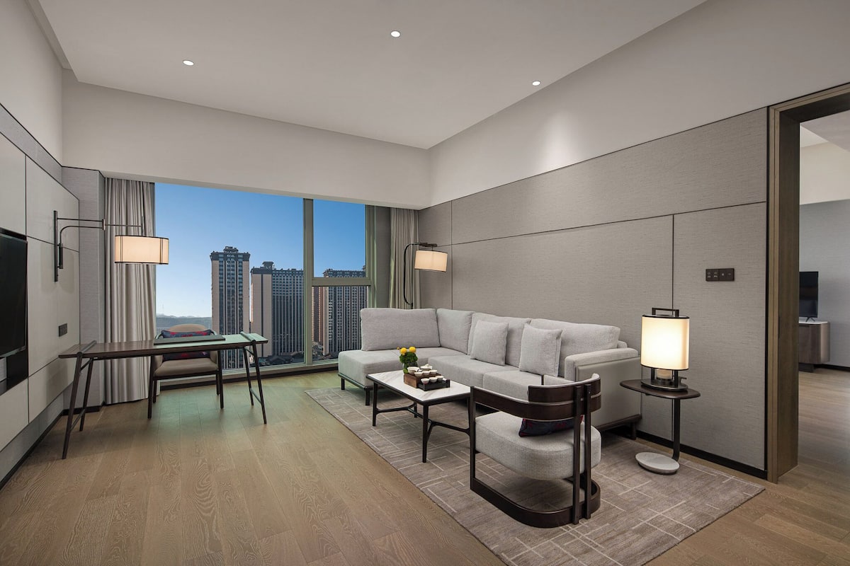 A modern hotel room with a large window, a sofa, a coffee table, a TV, and a lamp.